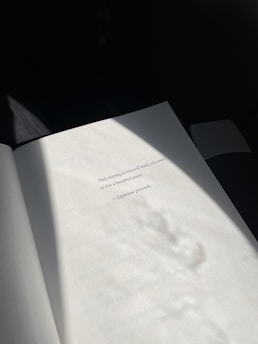 An open book page is illuminated by soft, natural light, revealing a printed quote attributed to a Japanese proverb. The shadows cast create a tranquil and contemplative atmosphere.