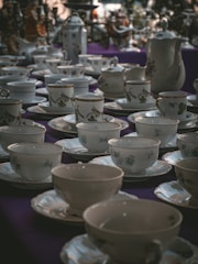 A collection of ornate teacups and saucers displayed on a table. The ceramics feature delicate floral designs and are arranged neatly across a dark purple tablecloth. At the back, there are teapots and other tableware, adding to the elegant and vintage ambiance of the scene.