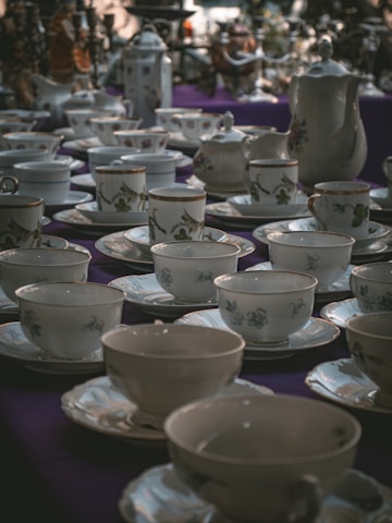 A collection of ornate teacups and saucers displayed on a table. The ceramics feature delicate floral designs and are arranged neatly across a dark purple tablecloth. At the back, there are teapots and other tableware, adding to the elegant and vintage ambiance of the scene.