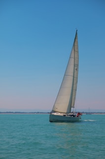 A sailboat gliding across clear turquoise waters under a bright blue sky. The sail is prominently hoisted, catching the breeze, while passengers are visible on the deck. In the distance, a faint coastline is seen along with another small vessel.