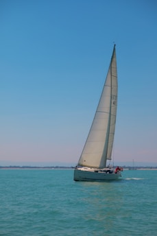 A sailboat gliding across clear turquoise waters under a bright blue sky. The sail is prominently hoisted, catching the breeze, while passengers are visible on the deck. In the distance, a faint coastline is seen along with another small vessel.