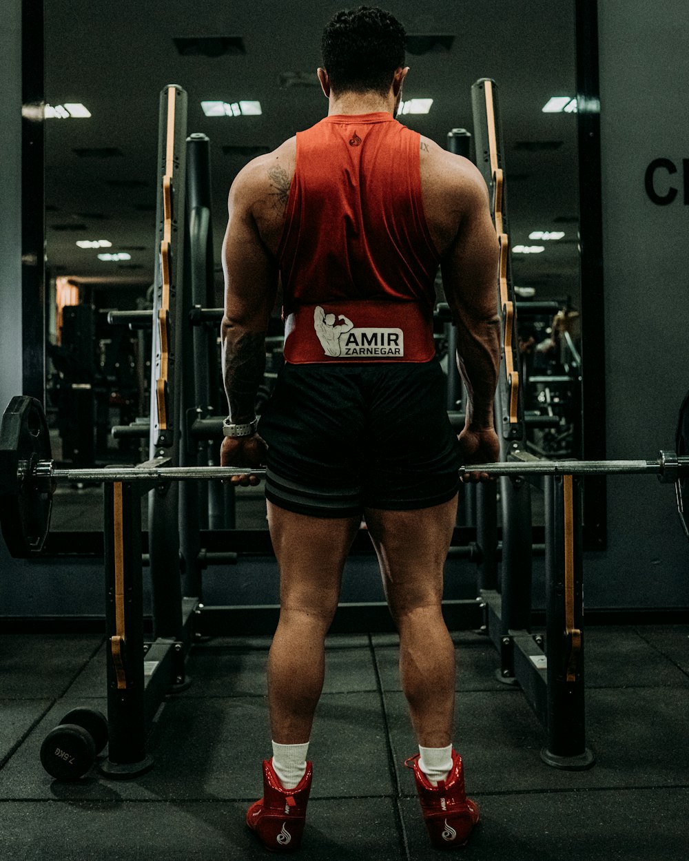 a man in a red shirt and black shorts standing in front of a barbell