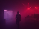 A dimly lit room with red and purple lighting creates a moody atmosphere. Silhouettes of people and electronic equipment are visible against the background, with hazy smoke or fog adding to the mysterious ambiance.