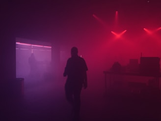 A dimly lit room with red and purple lighting creates a moody atmosphere. Silhouettes of people and electronic equipment are visible against the background, with hazy smoke or fog adding to the mysterious ambiance.