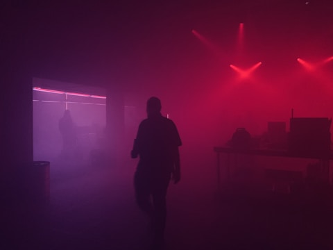 A dimly lit room with red and purple lighting creates a moody atmosphere. Silhouettes of people and electronic equipment are visible against the background, with hazy smoke or fog adding to the mysterious ambiance.