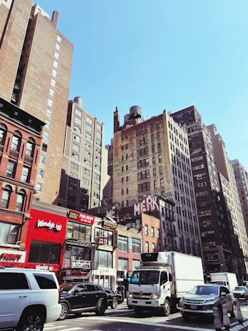 Tall buildings line the street, featuring a mix of older brick structures and modern architecture. The foreground includes several vehicles such as a truck, SUV, and car navigating a busy urban street. Various signs, including one for Wendy's and a 'FOR RENT' sign, add color and text to the scene. A clear blue sky enhances the cityscape.