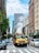 A bright yellow school bus travels down a busy city street between tall buildings. Steam rises from a vent in the road, creating a prominent white cloud. Numerous cars and a cyclist move alongside the bus, which has the number 148 on its front. The scene includes a mix of modern and older architectural styles, with clear blue skies overhead.