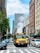 A bright yellow school bus travels down a busy city street between tall buildings. Steam rises from a vent in the road, creating a prominent white cloud. Numerous cars and a cyclist move alongside the bus, which has the number 148 on its front. The scene includes a mix of modern and older architectural styles, with clear blue skies overhead.