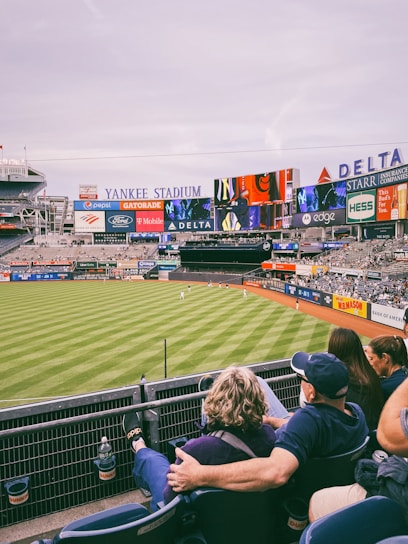 A baseball stadium filled with spectators watching a game on a neatly maintained grass field. The stadium features large advertising screens displaying various logos and brands. Fans sitting in the foreground are dressed casually, enjoying the sporting event.