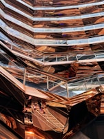 An intricate, modern architectural structure with multiple levels and glass railings, featuring a metallic copper finish that creates reflections and varying patterns. The design has a honeycomb-like appearance with angular, geometric shapes dominating the composition.