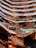 An intricate, modern architectural structure with multiple levels and glass railings, featuring a metallic copper finish that creates reflections and varying patterns. The design has a honeycomb-like appearance with angular, geometric shapes dominating the composition.