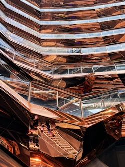 An intricate, modern architectural structure with multiple levels and glass railings, featuring a metallic copper finish that creates reflections and varying patterns. The design has a honeycomb-like appearance with angular, geometric shapes dominating the composition.