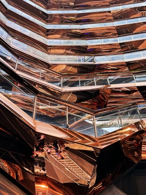 An intricate, modern architectural structure with multiple levels and glass railings, featuring a metallic copper finish that creates reflections and varying patterns. The design has a honeycomb-like appearance with angular, geometric shapes dominating the composition.