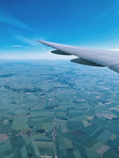 An aerial view from an airplane window showcasing a patchwork of agricultural fields below, with varying shades of green and brown. The airplane wing is visible in the foreground against a clear blue sky.