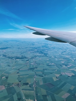 An aerial view from an airplane window showcasing a patchwork of agricultural fields below, with varying shades of green and brown. The airplane wing is visible in the foreground against a clear blue sky.