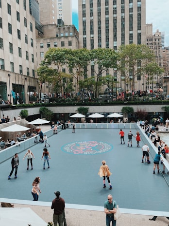 People are roller skating on an urban skating rink surrounded by tall buildings. There are several skaters of various ages on the rink, while others watch from the sidelines. Trees and umbrellas provide shade, and the area is bustling with activity.