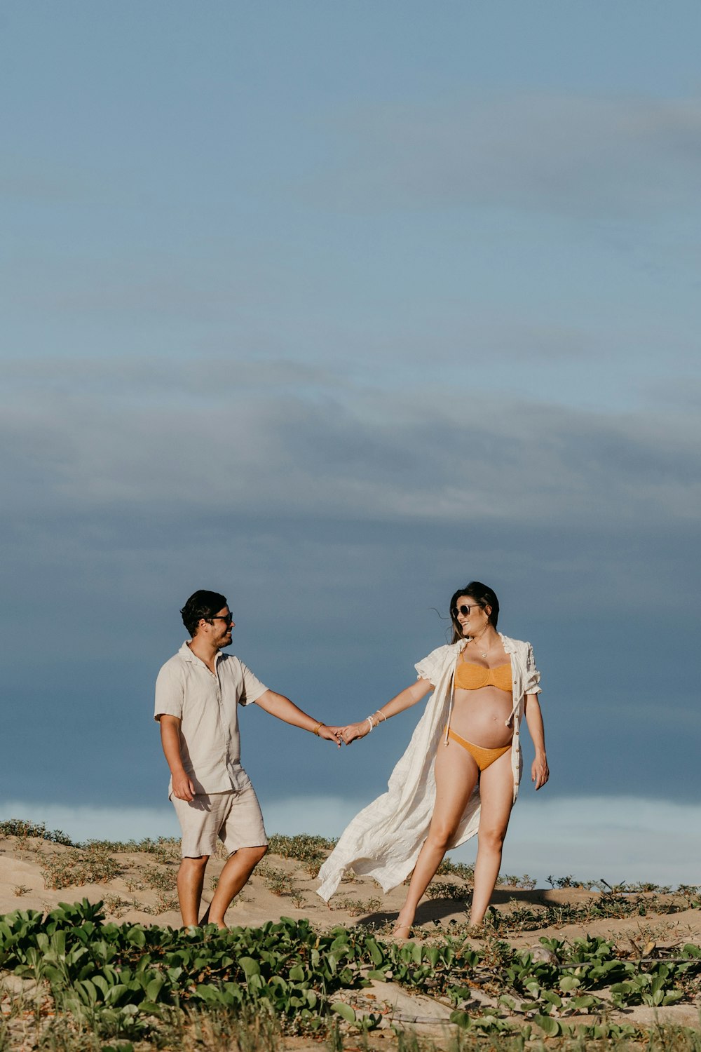 a pregnant woman in a yellow bikini and a man in a white shirt hold hands