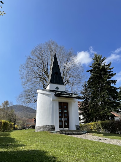 A small white chapel with a pointed dark roof stands in a green grassy area, surrounded by trees. The sky is bright blue and clear, with a few scattered clouds. A path leads to the chapel, and there are hills visible in the background.