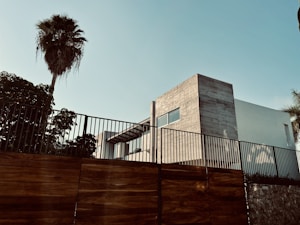 A modern architectural building with clean lines and a minimalist design is enclosed by a tall metal and wooden fence. A large palm tree stands prominently to the left, providing a contrast between nature and urban structure. The sky is clear, contributing to a serene atmosphere.