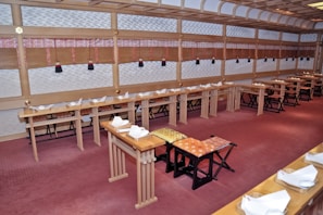 An empty room with minimalist traditional Japanese decor features rows of low wooden tables lined with ornate place settings and folded napkins. The walls are adorned with bamboo blinds and patterned panels. The carpet covers the floor in a soft red hue, while the ceiling showcases wooden beams.