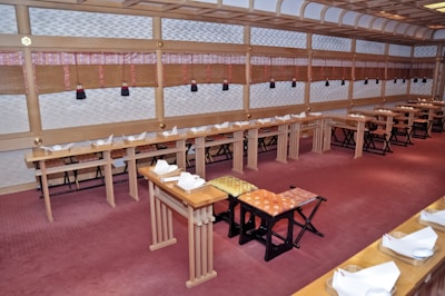 An empty room with minimalist traditional Japanese decor features rows of low wooden tables lined with ornate place settings and folded napkins. The walls are adorned with bamboo blinds and patterned panels. The carpet covers the floor in a soft red hue, while the ceiling showcases wooden beams.