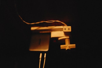 A dimly lit scene featuring a group of electronic devices and cables. There is an electrical outlet, a power strip, a smartphone, and connected earphones. The composition is slightly abstract with a focus on the interplay of shadows and angles.