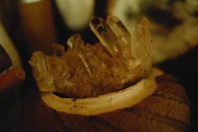 Clusters of translucent crystal formations rest atop a textured brown surface. The lighting is warm and subdued, giving the crystals an amber hue. The background is softly blurred, focusing attention on the crystal details.