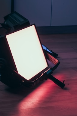 A large, rectangular LED light panel lies on a wooden floor, emitting a bright pinkish-red glow. The dark metal frame surrounds the panel, and the light reflects off the floor, enhancing the color.