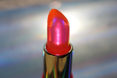 A lipstick with a glossy, holographic finish is featured, showcasing a vibrant red and orange hue. The metallic casing highlights the lipstick's sleek design. Light reflects off the surface, creating a shimmering effect.