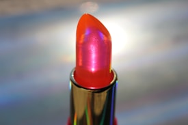 A lipstick with a glossy, holographic finish is featured, showcasing a vibrant red and orange hue. The metallic casing highlights the lipstick's sleek design. Light reflects off the surface, creating a shimmering effect.