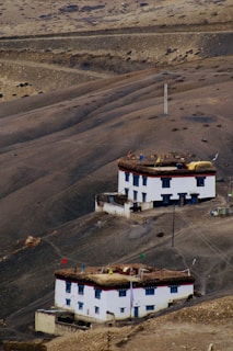 A barren mountainous landscape features two traditional white buildings with flat roofs, adorned with colorful prayer flags and set against a backdrop of dry, rolling hills. The scenery conveys a sense of rugged isolation and natural beauty.