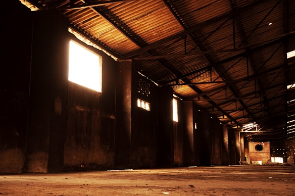 An expansive, empty warehouse interior with a high, slanted roof made of corrugated metal panels. Light streams in through several large rectangular windows, casting warm, golden tones across the walls and floor. The structure forms an elongated space with visible support beams and a vacant expanse of concrete flooring.