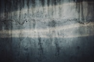 A textured concrete wall with visible cracks and water stains. The surface has dark and light streaks running horizontally, showing signs of wear and age.