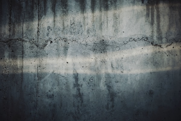 A textured concrete wall with visible cracks and water stains. The surface has dark and light streaks running horizontally, showing signs of wear and age.