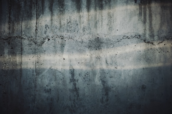 A textured concrete wall with visible cracks and water stains. The surface has dark and light streaks running horizontally, showing signs of wear and age.