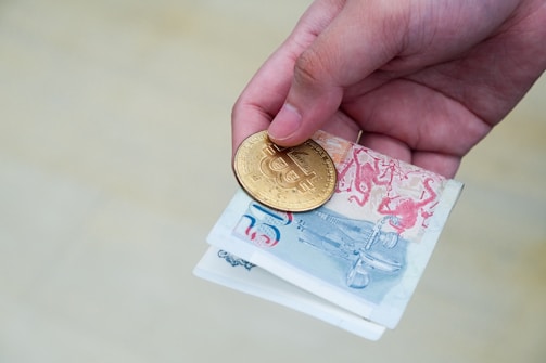A hand is holding a gold Bitcoin coin and a few paper banknotes. The background is plain and out of focus.