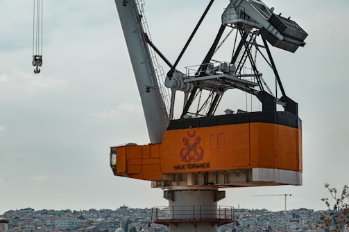 A large industrial crane with an orange base and complex metal framework towers above an urban landscape filled with densely packed buildings. The sky is overcast, giving the scene a muted lighting effect. The name 'Haliç Tersanesi' is prominently displayed on the crane.