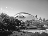 An architectural structure with a large, arched design stands prominently against a backdrop of modern buildings. Lush greenery with palm trees and bushes surrounds the foreground, and people can be seen cycling along a path. The scene is captured in black and white, giving it a timeless quality.
