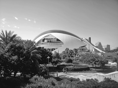 An architectural structure with a large, arched design stands prominently against a backdrop of modern buildings. Lush greenery with palm trees and bushes surrounds the foreground, and people can be seen cycling along a path. The scene is captured in black and white, giving it a timeless quality.