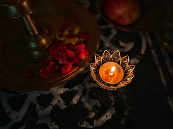 A burning candle in an ornate holder sits next to a brass tray adorned with red and yellow flower petals. The setting is dimly lit, creating a warm and serene ambiance.