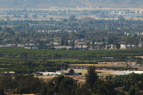 A vast landscape view of a rural area with extensive agricultural fields and scattered buildings. Trees and greenery dominate the foreground, while hills and small settlements are visible in the distance. A network of roads and utility lines intersect the landscape, contributing to a sense of human development amidst natural surroundings.