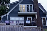 A two-story house with dark blue siding and a brick chimney features multiple signs with social and political messages displayed on its porch and windows. The house is bordered by a wooden fence, and there are trees surrounding the structure.
