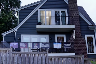 A two-story house with dark blue siding and a brick chimney features multiple signs with social and political messages displayed on its porch and windows. The house is bordered by a wooden fence, and there are trees surrounding the structure.