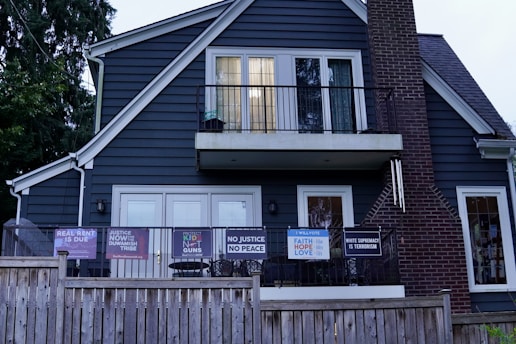 A two-story house with dark blue siding and a brick chimney features multiple signs with social and political messages displayed on its porch and windows. The house is bordered by a wooden fence, and there are trees surrounding the structure.