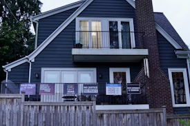 A two-story house with dark blue siding and a brick chimney features multiple signs with social and political messages displayed on its porch and windows. The house is bordered by a wooden fence, and there are trees surrounding the structure.