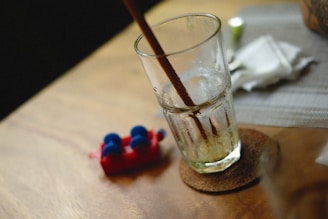 A partially filled glass with what seems to be ice and remnants of a drink is placed on a cork coaster on a wooden surface. A brown straw is inserted in the glass. To the left, a small red object with blue elements is visible, resembling a toy or small apparatus. In the background, a white napkin and a small container are placed on a gray placemat.