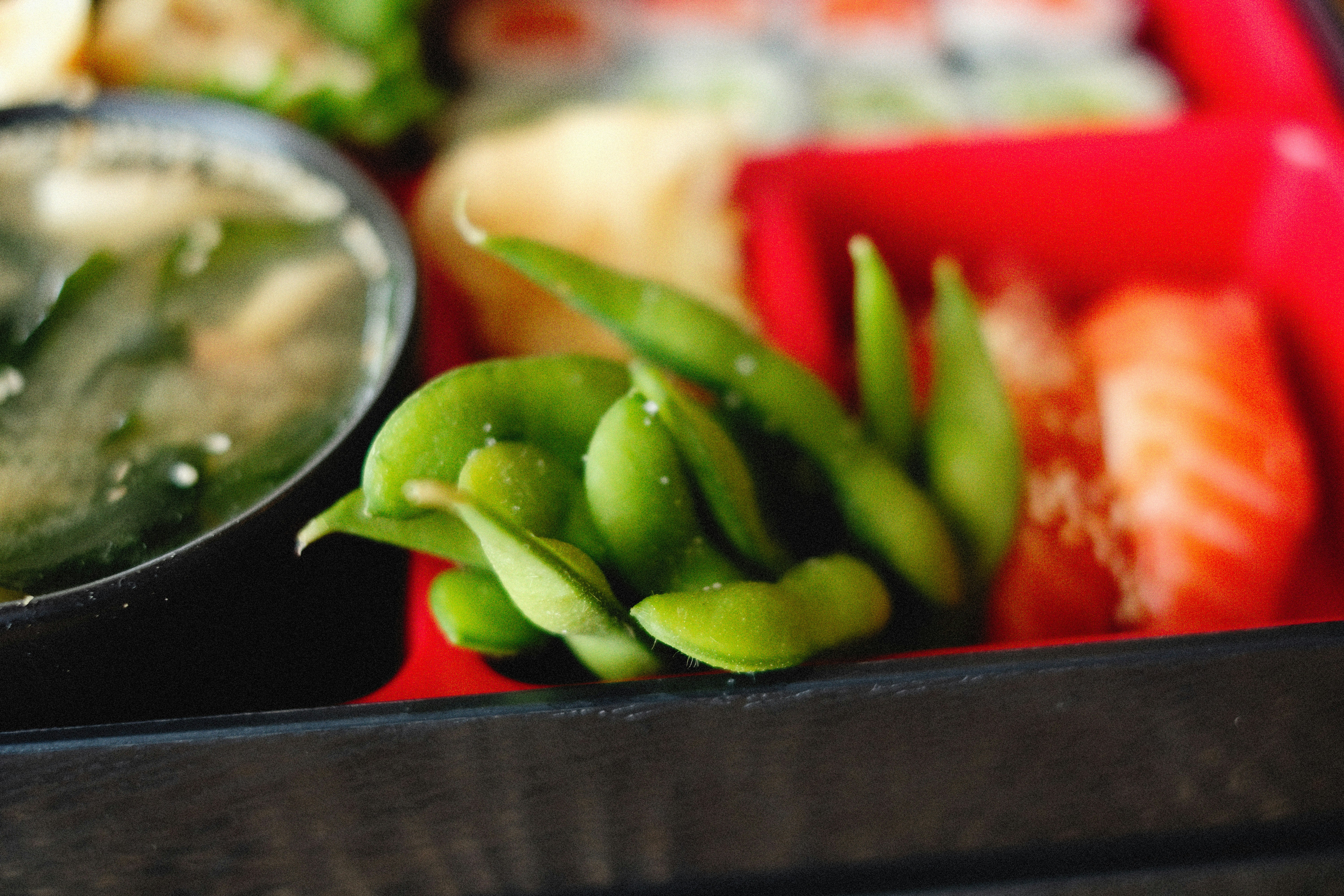 A close-up of a bento box containing a variety of food items. Edamame pods sit in the center, surrounded by sushi rolls partially visible in the background, a bowl of miso soup on the left, and a slice of salmon sashimi on the right. The tray is sectioned into bright red compartments.