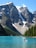 A stunning mountain landscape with towering, rugged peaks partially covered in snow. A turquoise lake stretches across the foreground, reflecting the clear blue sky. A canoe with two people paddling is visible on the water, surrounded by dense evergreen forests.