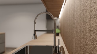 A modern kitchen faucet with a flexible coil spring design is mounted on a smooth wooden countertop. There is a sleek black sink beneath it, and the setup is illuminated by a strip of light attached to the underside of a wooden shelf above. The background features light-colored textured walls and a small plant.