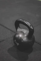 A matte black kettlebell casting a sharp shadow on a textured dark surface.